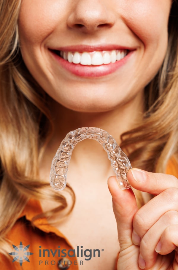 Your Path to a Straighter Smile Starts Here. A woman is smiling and holding Invisalign aligners in her hands